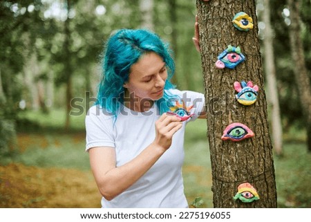 Young woman with turquoise dyed hair in white T shirt considering handmade colorful eye amulet, green park background, female art handmade artist at outdoor art exhibition