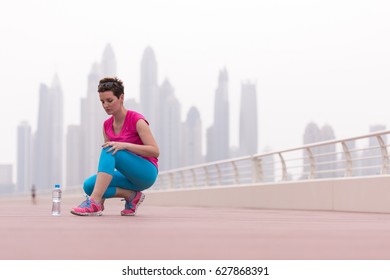 Young Woman Tying Shoelaces On Sneakers On A Promenade With A Big City In The Background. Standing Next To A Bottle Of Water. Exercise Outdoors