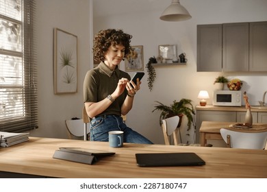 Young woman typing message on mobile phone while sitting in the kitchen and drinking coffee - Powered by Shutterstock