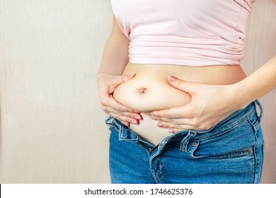 A young woman in unbuttoned trousers holds hands, squeezes belly fat at the waistline on jeans on light background, copy space. The concept of overweight, weight loss, diet, obesity, junk food.