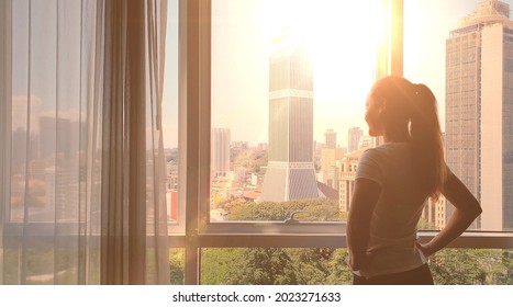 Young Woman Uncover The Big Window And Looking Out Her Apartment On The City Buildings. Sunrise In The City.