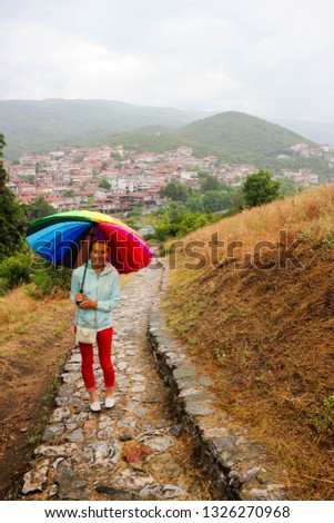 Young woman under the colorful umbrella in rainy weather on the stone path with view to the city and olympic mountains on the background, Greece 