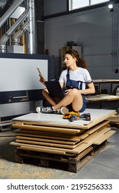 Young Woman In Uniform Sits On A Stack Of Chipboard Slabs With Laptop And Paper Blueprint In Hand