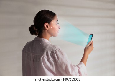 Young woman unlocking smartphone with facial scanner near white wall. Biometric verification