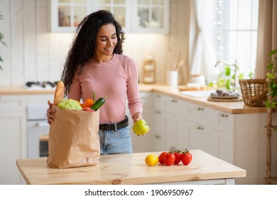 Young Woman Unpacking Paper Bag With Fresh Vegetables And Fruits After Grocery Shopping, Happy Brunette Lady Enjoying Organic Healthy Food, Smiling Female Standing In Cozy Kitchen At Home, Copy Space - Powered by Shutterstock