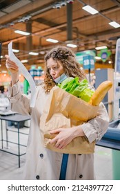 Young Woman Is Unpleasantly Surprised While Looking Into A Paper Check In A Grocery Supermarket And Holding A Shopping Bag. Rising Food Prices.