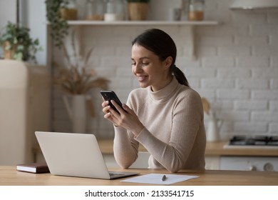 Young woman use cellphone sit at table in kitchen share text messages in social media, enjoy new vlog, watch internet content take break from work on laptop. Tech, e-services, mobile app usage concept