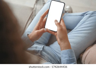 Young Woman User Customer Hold Smart Phone Mock Up White Screen In Hand Use Mobile Shopping App, Check Social Media News, Texting Mobile Sms Order Food Delivery Sit On Sofa At Home. Over Shoulder View