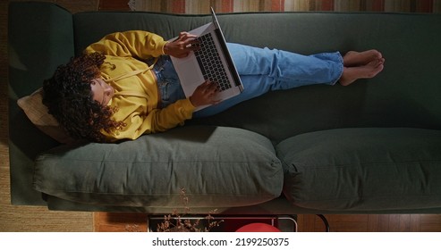 A Young Woman Uses Her Laptop While Sitting On Comfortable Sofa. A Black Hispanic Girl Accessing Social Media On Computer. From Above Angle Of Person Browsing Internet On Computer On Couch At Night