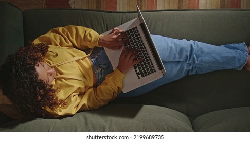 A Young Woman Uses Her Laptop While Sitting On Comfortable Sofa. A Black Hispanic Girl Accessing Social Media On Computer. From Above Angle Of Person Browsing Internet On Computer On Couch At Night