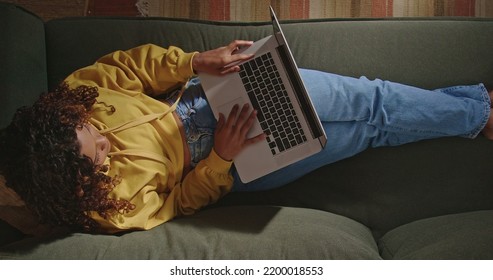 A Young Woman Uses Her Laptop While Sitting On Comfortable Sofa. A Black Hispanic Girl Accessing Social Media On Computer. From Above Angle Of Person Browsing Internet On Computer On Couch At Night