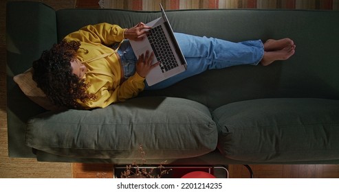 A Young Woman Uses Her Laptop While Sitting On Comfortable Sofa. A Black Hispanic Girl Accessing Social Media On Computer. From Above Angle Of Person Browsing Internet On Computer On Couch At Night