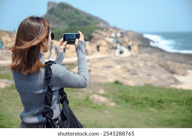 A young woman uses her smartphone to take a picture of the beautiful coastal scenery at Yehliu Geopark in Taiwan. Taking in the beauty of nature while fusing themes of tourism, adventure, and travel - Powered by Shutterstock