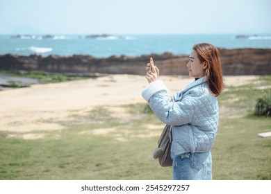 A young woman uses her smartphone to take a picture of the beautiful coastal scenery at Yehliu Geopark in Taiwan. Taking in the beauty of nature while fusing themes of tourism, adventure, and travel - Powered by Shutterstock