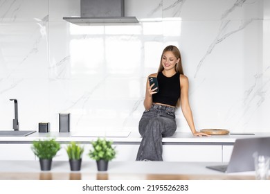 Young woman uses smartphone in the kitchen with laptop on the countertop. - Powered by Shutterstock