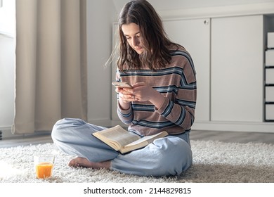A young woman uses a smartphone while sitting with a book in her hands. - Shutterstock ID 2144829815