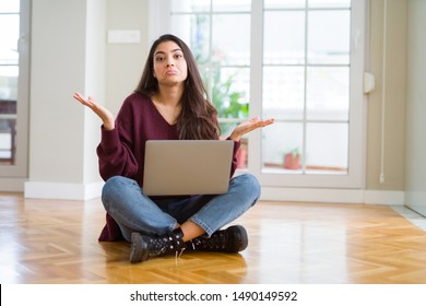 Young Woman Using Computer Laptop Sitting On The Floor Clueless And Confused Expression With Arms And Hands Raised. Doubt Concept.