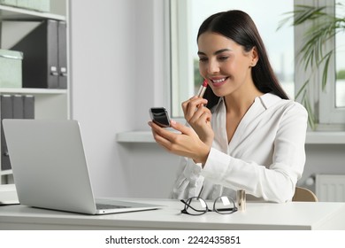 Young woman using cosmetic pocket mirror while applying makeup in office - Powered by Shutterstock