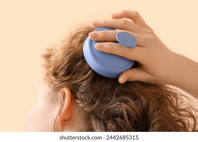 Young woman using hair scalp massager on beige background, closeup