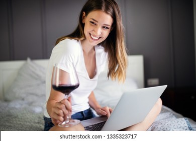 Young woman using her laptop while relaxing on bed and drinking wine