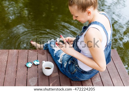 Young woman using her smartphone sitting on the pier and drinking hot coffee