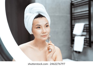 Young Woman Using Jade Roller In Modern Bathroom: A serene Asian woman with a towel on her head uses a jade facial roller, exuding calm and self-care in a stylish bathroom setting. - Powered by Shutterstock