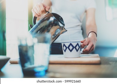 Young Woman Using Kettle For Make Tea Or Black Coffee On Kitchen At Room Interior Background.Women's Hands Pour Water From A Teapot Into A Cup. Blurred, Flares Effect