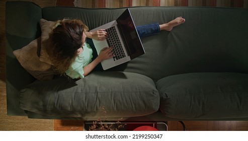 Young Woman Using Laptop Computer On Couch. From Above Angle Of Millennial Girl Browsing Internet Online With Modern Technology Device. Girl Studying In The Evening At Home Living Room