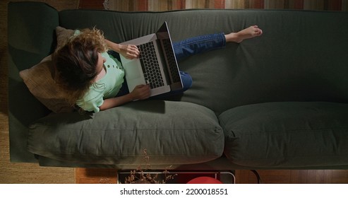 Young Woman Using Laptop Computer On Couch. From Above Angle Of Millennial Girl Browsing Internet Online With Modern Technology Device. Girl Studying In The Evening At Home Living Room