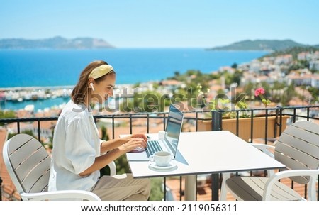Young woman using laptop computer at cafe balcony of resort hotel with sea view, working typing emails browsing online enjoying drinking coffee