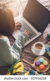 Young Woman Using Laptop At Cozy Home Atmosphere On The Bed With Cup Of Coffee And Macaroon.