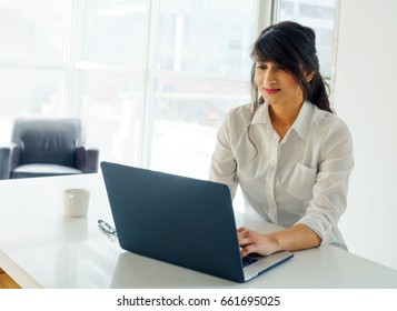 Young woman using laptop at desk, day, with window behind. young adult arabic pretty 20 years old