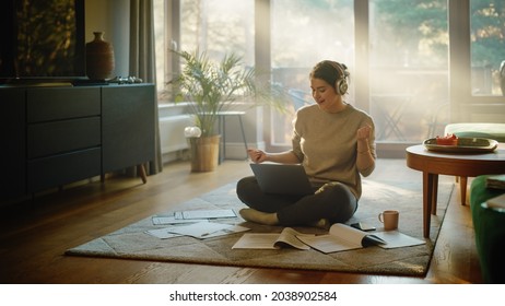 Young Woman Using Laptop at Home, Does Remote Work, Listens Music through Headphones. Beautiful Smiling Girl Sitting on the Floor Does Research on Papers, Documents, Brainstorms Creative Project - Powered by Shutterstock