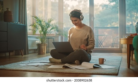 Young Woman Using Laptop at Home, Does Remote Work, Listens Music through Headphones. Beautiful Smiling Girl Sitting on the Floor Does Research on Papers, Documents, Brainstorms Creative Project - Powered by Shutterstock