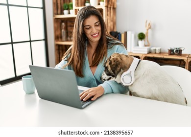 Young woman using laptop sitting on table with dog wearing headphones at home