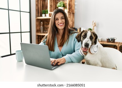 Young woman using laptop sitting on table with dog wearing headphones at home
