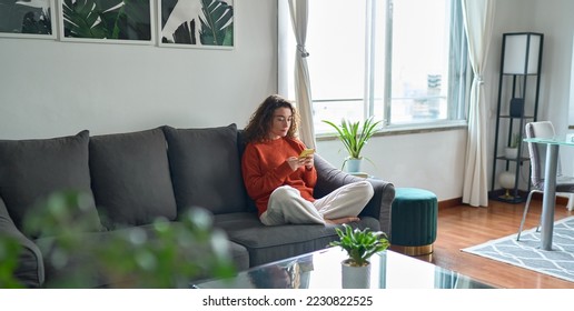 Young woman using mobile cell phone sitting on couch at home. Pretty relaxed lady holding smartphone buying in ecommerce shop, watching videos online, texting or checking social media news. - Powered by Shutterstock