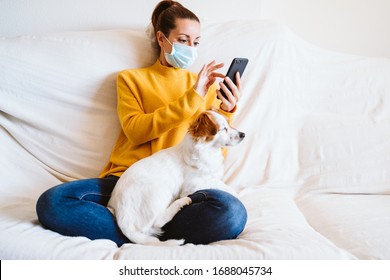young woman using mobile phone, cute small dog besides. Sitting on the couch, wearing protective mask. Stay home concept during coronavirus covid-2019