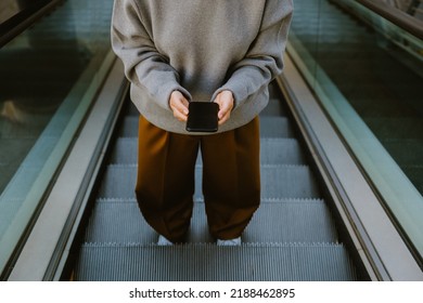 Young Woman Using Mobile Phone While Going Up The Escalator Outdoors