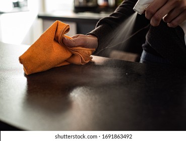Young woman using orange microfibre cloth and spray bottle to clean/sanitise dark worktop surface