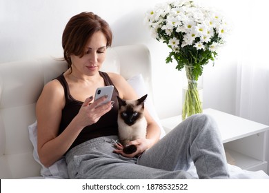 Young Woman Using Phone With Her Siamese Cat On Bedroom With Flowers.