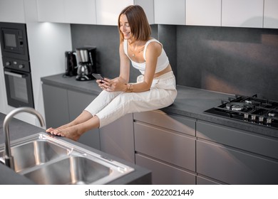 Young Woman Using Smart Phone While Sitting Relaxed At Modern Kitchen At Home. Concept Of Smart Kitchen Appliances And Cozy Home