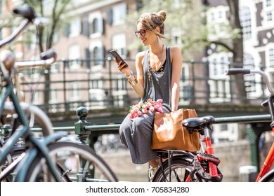 Young Woman Using A Smart Phone Sitting With Bag And Flowers Near The Water Channel In Amsterdam Old City