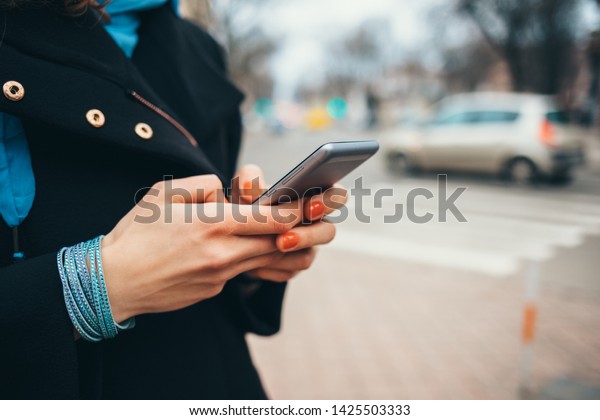 Young woman using smart phone\
outdoors holding it in her hands, close-up. Female wearing black\
coat typing text messages on mobile device on street near road with\
cars.