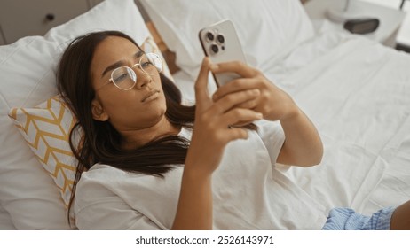 Young woman using smartphone in bedroom lying on bed wearing glasses with brunette hair in a home interior setting - Powered by Shutterstock