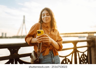 Young woman using smartphone by the waterfront during sunset, wearing a casual outfit while enjoying the warm evening light. Lifestyle, travel, nature, active life. - Powered by Shutterstock