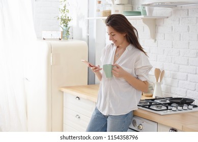 Young woman using smartphone leaning at kitchen table with coffee mug and organizer in a modern home. Smiling woman reading phone message. Brunette happy girl typing a text message - Powered by Shutterstock