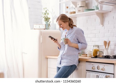 Young woman using smartphone leaning at kitchen table with coffee mug and organizer in a modern home. Smiling woman reading phone message. Brunette happy girl typing a text message