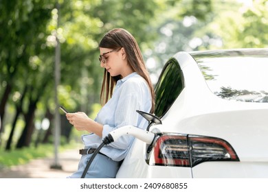 Young woman using smartphone online banking application to pay for electric car battery charging from EV charging station during vacation holiday road trip at national park or summer forest. Exalt - Powered by Shutterstock