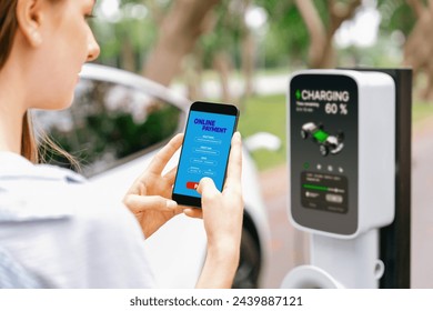 Young woman using smartphone online banking application to pay for electric car battery charging from EV charging station during vacation holiday road trip at national park or summer forest. Exalt - Powered by Shutterstock